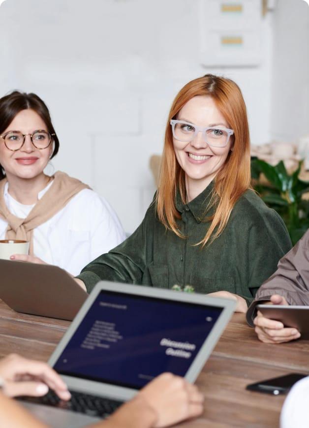 Two women smiling in an office, across from someone working on their laptop.